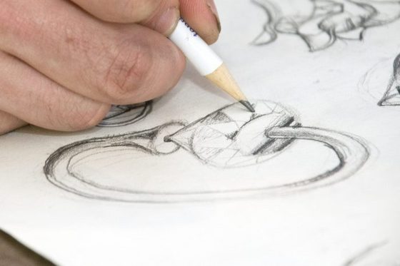 Detailaufnahme einer Hand, die einen Bleistift hält und auf einem weißen Papier Schmuckstücke skizziert.