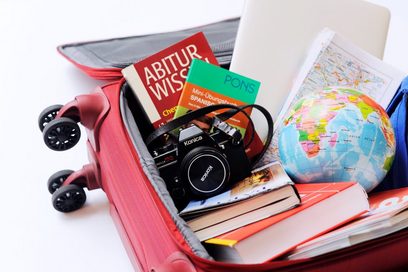 Blick in einen roten Trolle, in dem verschiedene Reiseutensilien sind wie etwa eine Kamera, Bücher, ein Laptop, T-Shirts.