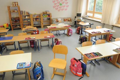 Blick in ein Klassenzimmer. Auf den Tischen liegen aufgeschlagene Federmäppchen, daneben stehen Schulranzen. An der rückwärtigen Wand des Raumes stehen Regale mit Arbeitsmaterialien und ein Sofa.