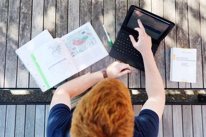 Junger Mann in blauem T-Shirt sitzt an einer Bank auf dem Campus einer Hochschule und arbeitet an einem Laptop.