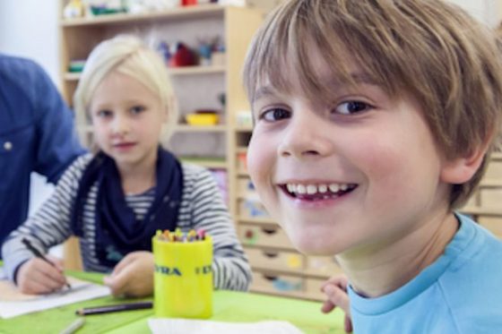 Ein kleiner Junge in blauem T-Shirt sitzt zusammen mit einem blonden Mädchen an einem großen Tisch, auf dem eine grüne Unterlage liegt. Die beiden Kinder malen mit Buntstiften auf Papier.