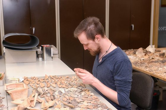 Ein Archäologe sitzt am Schreibtisch und begutachtet Fundstücke.