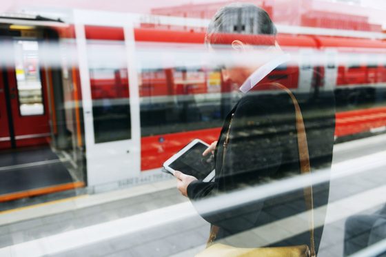 Ein Mann steht mit einem Tablet am Bahnsteig vor einem Zug, dessen Türen geöffnet sind.